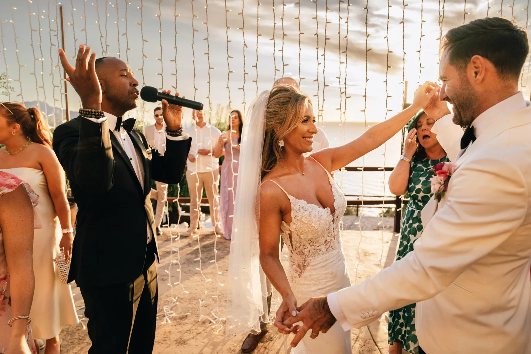 Hochzeitstanz mit Livegesang – Inan Lima, Hochzeitsband auf Mallorca Inan Lima singt live mit Mikrofon, während das Brautpaar im Sonnenuntergang unter Lichterketten am Meer auf Mallorca den Hochzeitstanz beginnt